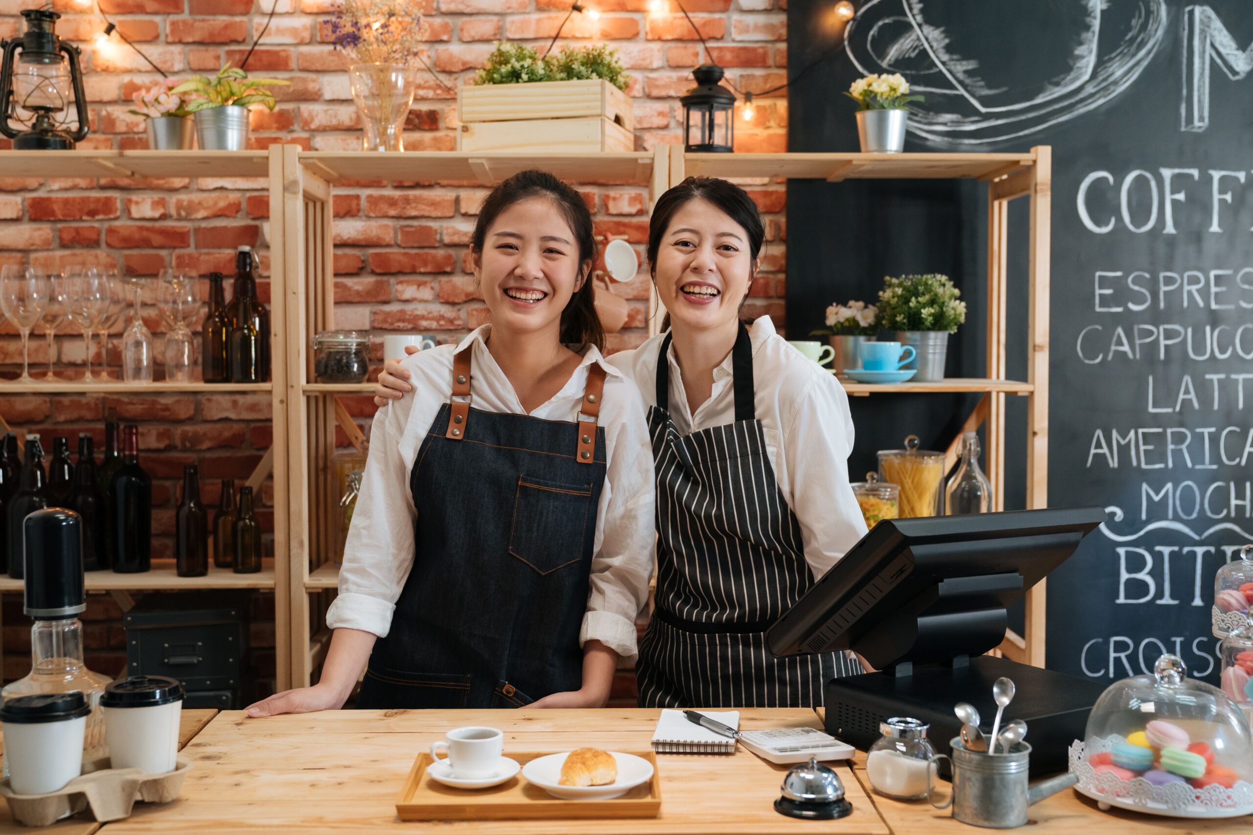 Two female baristas smile behind a POS terminal in a small business coffeehouse online-ready with Kinetic Business Internet