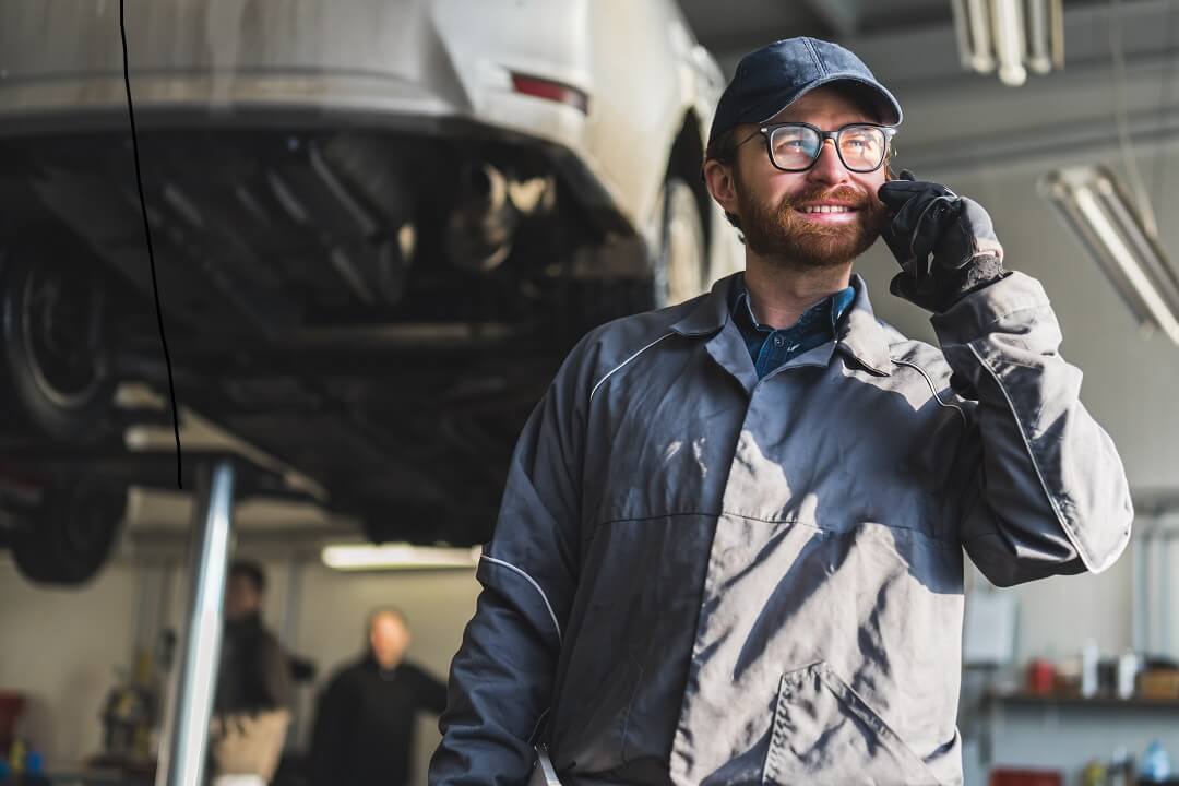 Male auto mechanic in coveralls using business phone while repairing a vehicle.