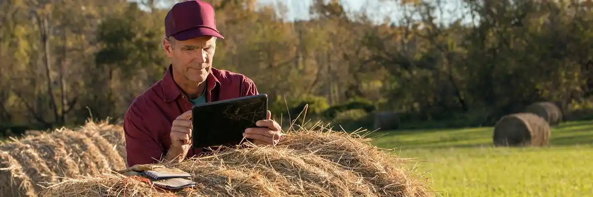 Farmer leans on hay bale and checks his wireless tablet device